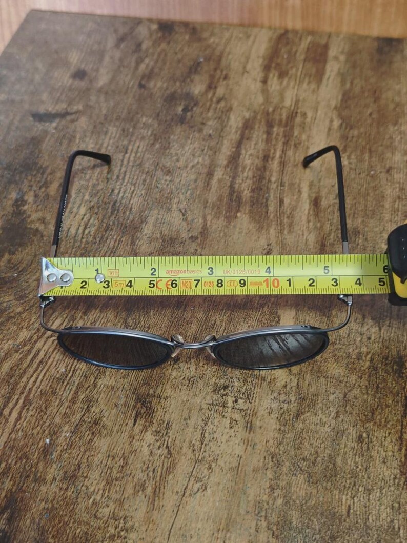May include: A pair of oval-shaped sunglasses with dark lenses and thin, dark metal frames. The glasses are measured with a tape measure, showing a length of approximately 5 inches.