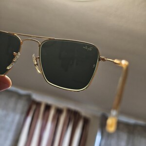 Puede incluir: Gafas de sol con montura dorada y lentes verde oscuro. Las gafas de sol tienen una forma cuadrada cl&aacute;sica y el logotipo de Ray-Ban es visible en la lente superior derecha. Las patillas son de color dorado claro.