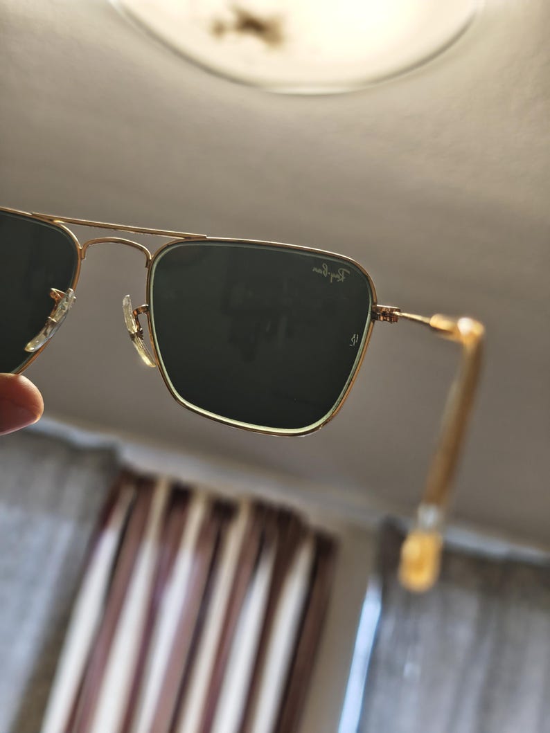 Puede incluir: Gafas de sol con montura dorada y lentes verde oscuro. Las gafas de sol tienen una forma cuadrada cl&aacute;sica y el logotipo de Ray-Ban es visible en la lente superior derecha. Las patillas son de color dorado claro.