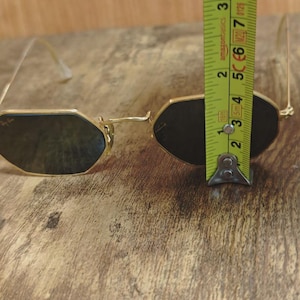 May include: Gold-framed sunglasses with dark lenses. The glasses have a unique octagonal shape. The image shows the glasses are approximately 2.5 inches in height. The glasses are on a wooden surface.