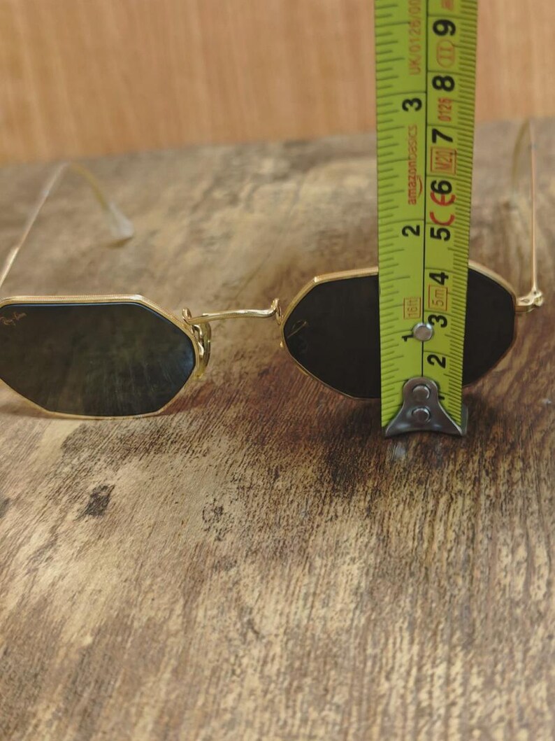 May include: Gold-framed sunglasses with dark lenses. The glasses have a unique octagonal shape. The image shows the glasses are approximately 2.5 inches in height. The glasses are on a wooden surface.