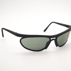 May include: Black sunglasses with a streamlined, curved design. The lenses are a muted green, set in a matte black frame. The arms curve down and back, giving a sporty and modern look.