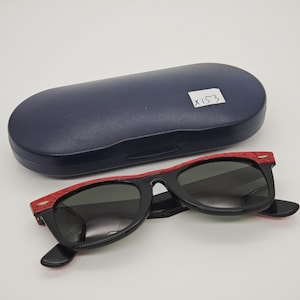 May include: Black sunglasses with red detailing and a dark blue case. The sunglasses feature a classic design with dark lenses and red frame accents. The case is oval-shaped. The text "X153" is visible on the case.