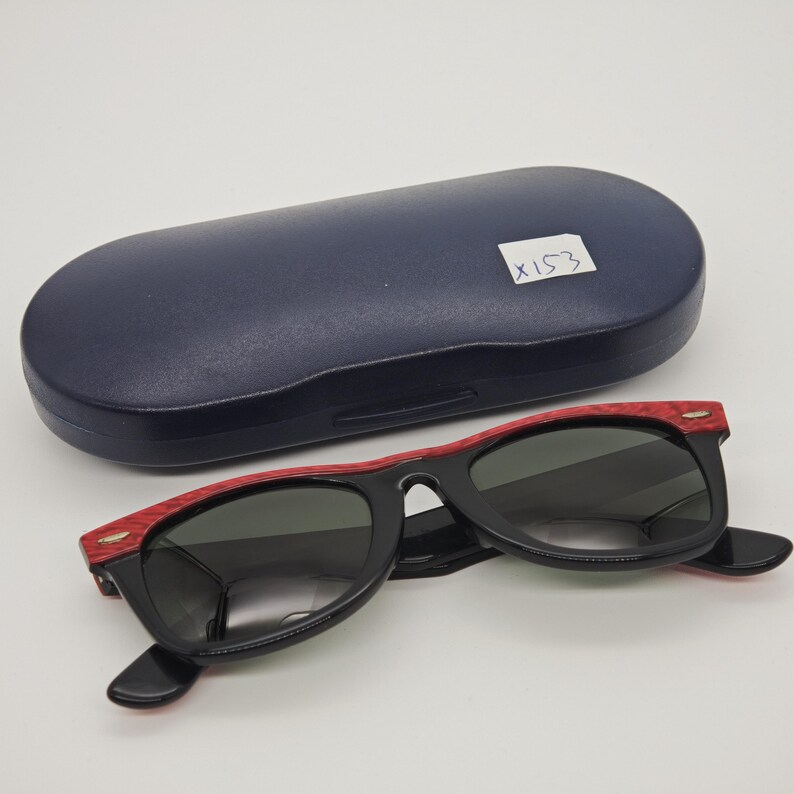 May include: Black sunglasses with red detailing and a dark blue case. The sunglasses feature a classic design with dark lenses and red frame accents. The case is oval-shaped. The text "X153" is visible on the case.