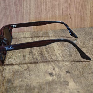 May include: Brown sunglasses with dark lenses. The frames have a tortoiseshell pattern and the Ray-Ban logo is visible on the left arm. The sunglasses are resting on a wooden surface.
