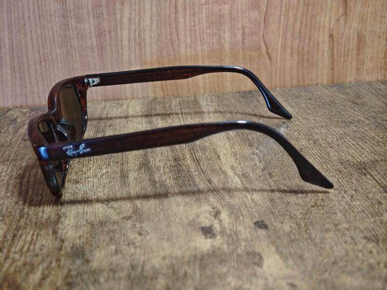 May include: Brown sunglasses with dark lenses. The frames have a tortoiseshell pattern and the Ray-Ban logo is visible on the left arm. The sunglasses are resting on a wooden surface.