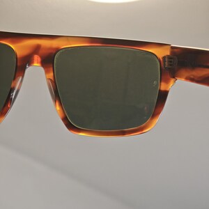 May include: Close-up of sunglasses with a tortoiseshell pattern frame and dark lenses. The sunglasses have a classic square shape and a thick frame. The lenses are a dark green color, and the frame has a warm, brown and amber color.