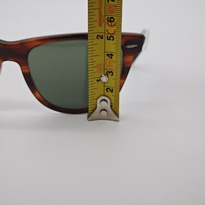 May include: Tortoiseshell sunglasses with green lenses. A yellow measuring tape is placed over the sunglasses, showing a measurement of approximately 2 inches. The sunglasses have a classic, retro design.
