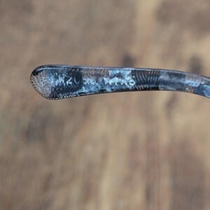May include: Close-up of a silver eyeglass arm. The arm is curved and has a textured, patterned design on the end. The metal has a slightly tarnished appearance, with areas of darker and lighter tones.