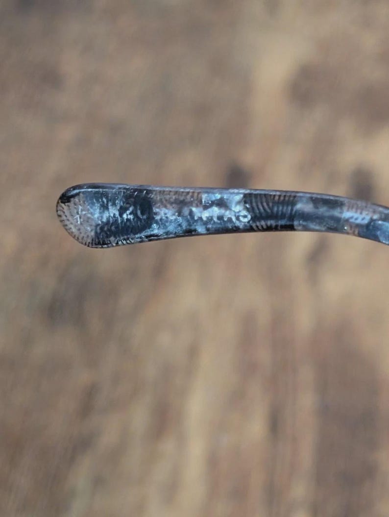 May include: Close-up of a silver eyeglass arm. The arm is curved and has a textured, patterned design on the end. The metal has a slightly tarnished appearance, with areas of darker and lighter tones.