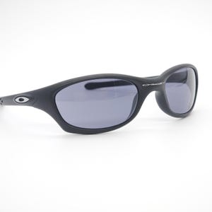 May include: Black Oakley sunglasses with gray lenses. The sunglasses have a curved frame and the Oakley logo is visible on the temples. The lenses are a dark gray color, and the frame is matte black.