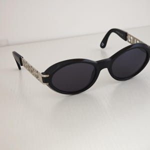 Puede incluir: Gafas de sol ovaladas con montura negra y lentes oscuras. Las patillas presentan detalles en metal plateado con un patr&oacute;n geom&eacute;trico repetitivo. Los extremos de las patillas son de pl&aacute;stico negro, con un dise&ntilde;o sencillo y funcional.