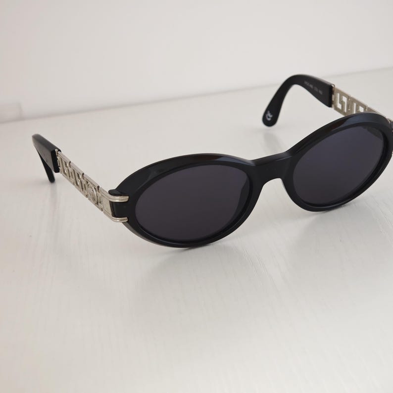 Puede incluir: Gafas de sol ovaladas con montura negra y lentes oscuras. Las patillas presentan detalles en metal plateado con un patr&oacute;n geom&eacute;trico repetitivo. Los extremos de las patillas son de pl&aacute;stico negro, con un dise&ntilde;o sencillo y funcional.