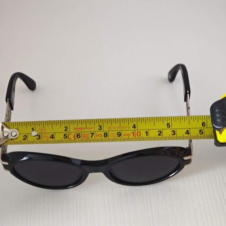 Puede incluir: Gafas de sol negras con monturas ovaladas y lentes oscuras. Una cinta m&eacute;trica amarilla est&aacute; colocada sobre la parte frontal de las monturas, mostrando un ancho de aproximadamente 14 cm desde el borde exterior de una bisagra a la otra.