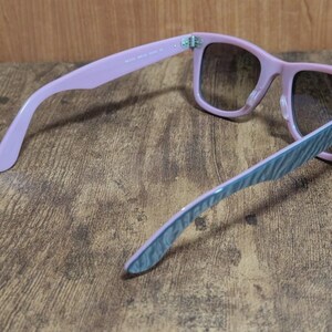 Puede incluir: Gafas de sol rosas con lentes degradadas. Las monturas son de color rosa suave y las patillas tienen un estampado de rayas azules y blancas. Las gafas de sol est&aacute;n sobre un fondo de madera.