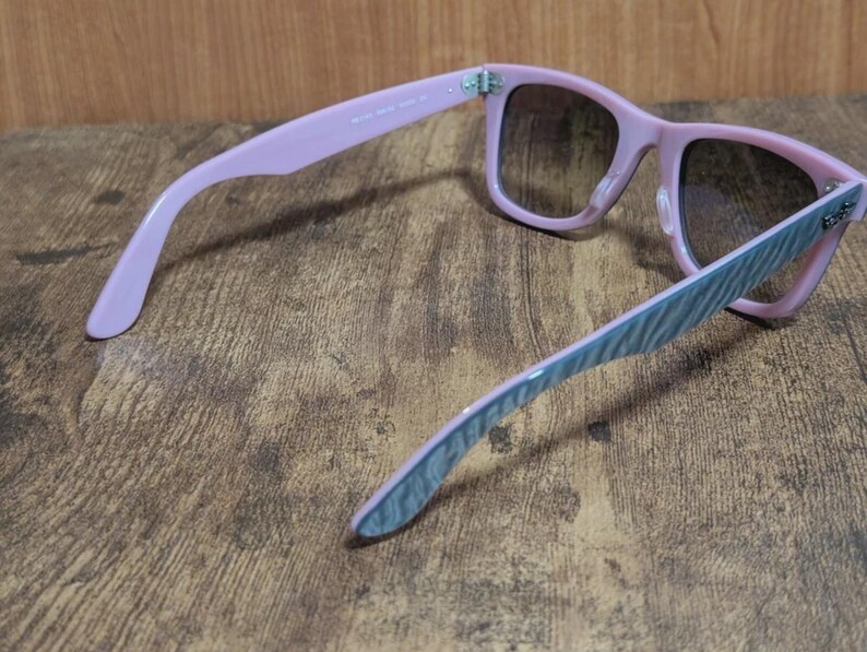 Puede incluir: Gafas de sol rosas con lentes degradadas. Las monturas son de color rosa suave y las patillas tienen un estampado de rayas azules y blancas. Las gafas de sol est&aacute;n sobre un fondo de madera.