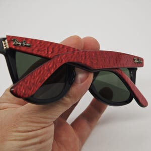 May include: Black sunglasses with red textured frames. The sunglasses have dark lenses and the Ray-Ban logo is visible on the frame. The sunglasses are held in a hand.