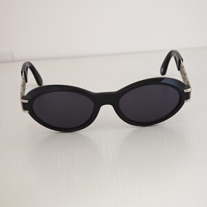 Puede incluir: Gafas de sol ovaladas con montura negra y lentes oscuras. Las patillas est&aacute;n formadas por una cadena de eslabones de metal plateado que se conecta a terminales negros curvados, creando un contraste definido con el frente negro s&oacute;lido.