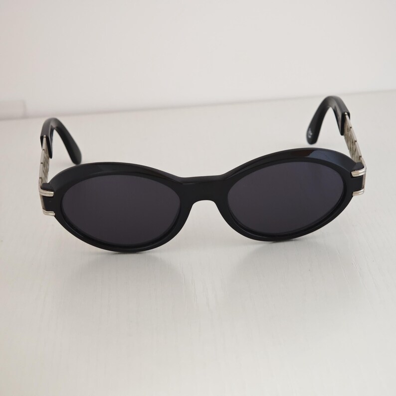 Puede incluir: Gafas de sol ovaladas con montura negra y lentes oscuras. Las patillas est&aacute;n formadas por una cadena de eslabones de metal plateado que se conecta a terminales negros curvados, creando un contraste definido con el frente negro s&oacute;lido.