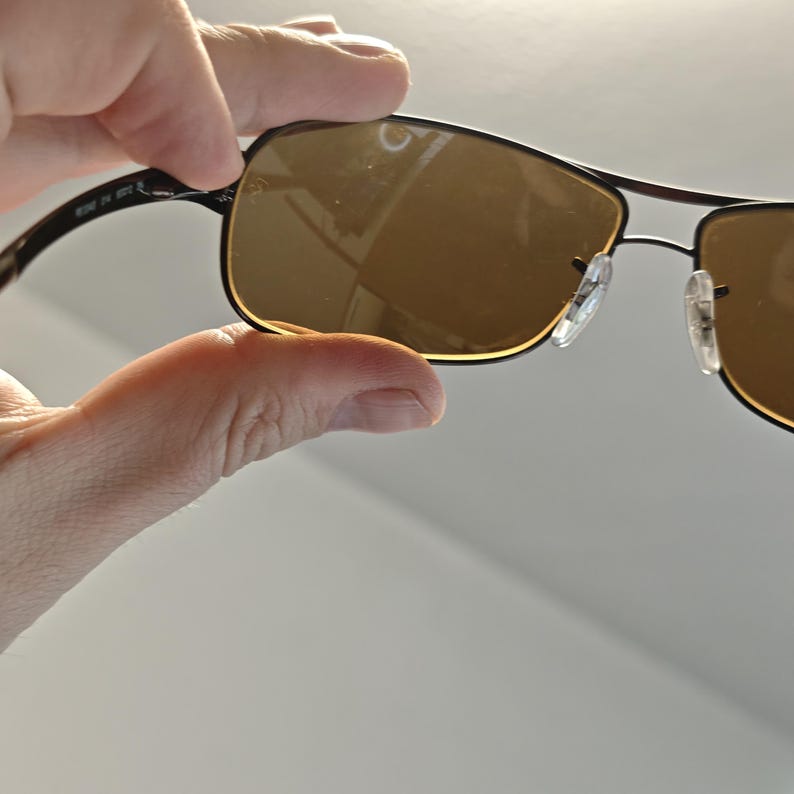 May include: Sunglasses with a dark brown metal frame and rectangular lenses. The lenses are a warm brown color. The sunglasses have a classic design with a bridge and nose pads.