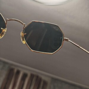May include: Gold-framed sunglasses with dark lenses. The sunglasses have an octagonal shape and a thin gold frame. The nose pads are a light tan color. The lenses are dark, and the arms are thin and gold.