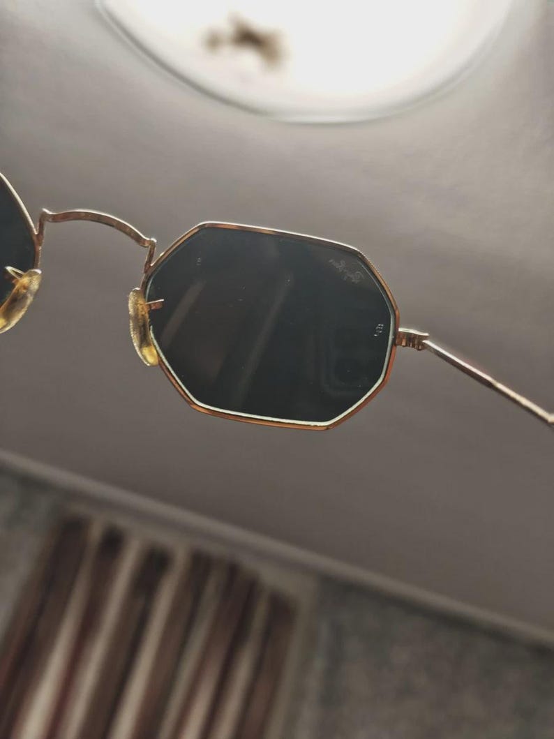 May include: Gold-framed sunglasses with dark lenses. The sunglasses have an octagonal shape and a thin gold frame. The nose pads are a light tan color. The lenses are dark, and the arms are thin and gold.
