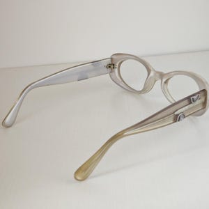 May include: A pair of eyeglasses with translucent, light beige frames and oval-shaped rims. The temples are a solid, opaque beige color and feature small, silver-toned metallic accents near the hinges, showing the back view of the eyewear.