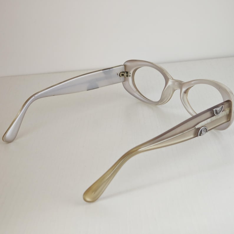May include: A pair of eyeglasses with translucent, light beige frames and oval-shaped rims. The temples are a solid, opaque beige color and feature small, silver-toned metallic accents near the hinges, showing the back view of the eyewear.