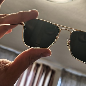 Puede incluir: Gafas de sol con montura dorada y lentes oscuras. Las gafas de sol tienen un dise&ntilde;o cl&aacute;sico de aviador con doble puente y lentes rectangulares. La montura es de color dorado brillante y las lentes son de color verde oscuro o negro.