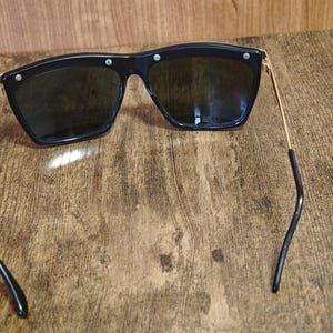 Peut inclure: Lunettes de soleil noires avec une monture g&eacute;om&eacute;trique et des verres fonc&eacute;s. Les lunettes ont des branches dor&eacute;es et de petits accents argent&eacute;s sur la monture. Les branches sont incurv&eacute;es aux extr&eacute;mit&eacute;s.