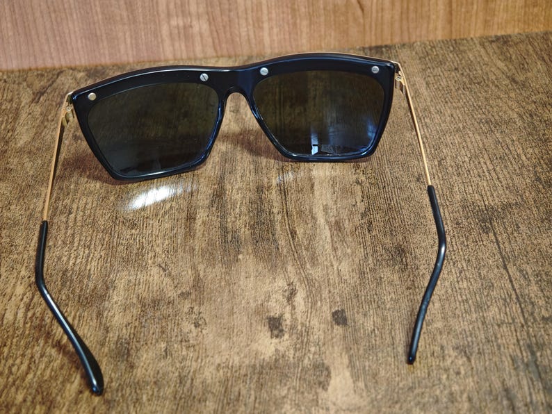 Peut inclure: Lunettes de soleil noires avec une monture g&eacute;om&eacute;trique et des verres fonc&eacute;s. Les lunettes ont des branches dor&eacute;es et de petits accents argent&eacute;s sur la monture. Les branches sont incurv&eacute;es aux extr&eacute;mit&eacute;s.