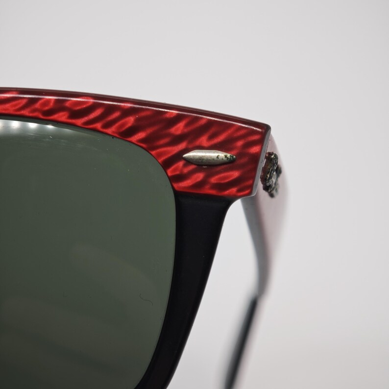 May include: Close-up of sunglasses with a red textured frame and dark lenses. The frame has a unique pattern and a silver accent near the hinge. The sunglasses are a classic style.
