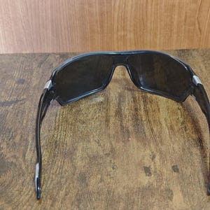 May include: Black sunglasses with dark lenses and a sporty design. The sunglasses have a thick frame and silver accents at the temples. The lenses are dark, providing protection from the sun.