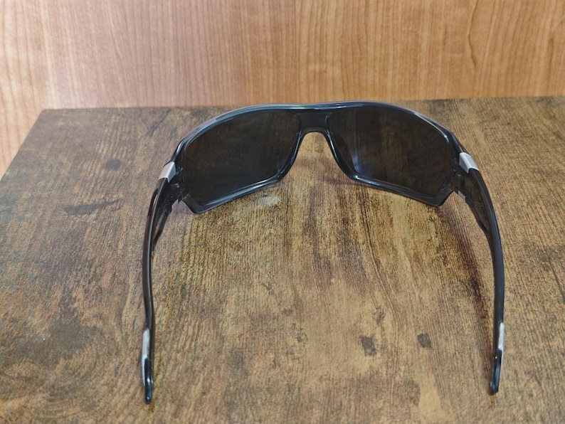 May include: Black sunglasses with dark lenses and a sporty design. The sunglasses have a thick frame and silver accents at the temples. The lenses are dark, providing protection from the sun.