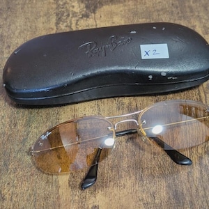 May include: A pair of Ray-Ban sunglasses with brown tinted lenses and a silver frame, resting on a wooden surface. The sunglasses are next to a black leather case with the Ray-Ban logo embossed on it. A white sticker with "X 2" is on the case.