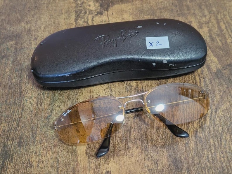 May include: A pair of Ray-Ban sunglasses with brown tinted lenses and a silver frame, resting on a wooden surface. The sunglasses are next to a black leather case with the Ray-Ban logo embossed on it. A white sticker with "X 2" is on the case.