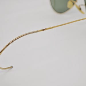 May include: Close-up of gold-toned sunglasses with green lenses. The frame shows signs of wear, with discoloration on the temple arm. The curved temple arm is visible, along with the hinge and lens.