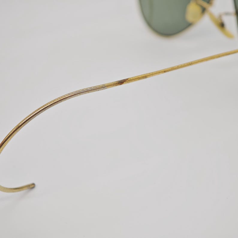May include: Close-up of gold-toned sunglasses with green lenses. The frame shows signs of wear, with discoloration on the temple arm. The curved temple arm is visible, along with the hinge and lens.