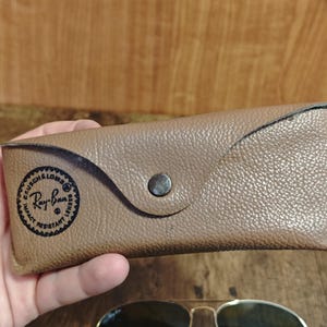 May include: A tan leather Ray-Ban sunglasses case with a snap closure. The case has a circular logo with the words "BAUSCH & LOMB" and "Ray-Ban" printed on it. The case is held in a hand.