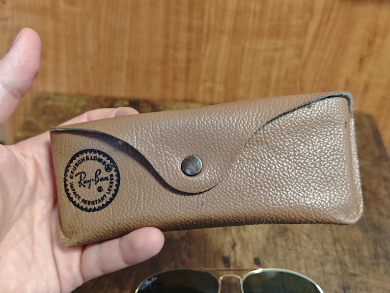 May include: A tan leather Ray-Ban sunglasses case with a snap closure. The case has a circular logo with the words "BAUSCH & LOMB" and "Ray-Ban" printed on it. The case is held in a hand.