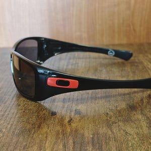 K&ouml;nnte beinhalten: Schwarze Sonnenbrille mit gl&auml;nzendem Finish und dunklen Gl&auml;sern. Die Sonnenbrille hat einen roten Akzent am Rahmen und ist vor einem Holzgrund platziert. Das Design ist sportlich und elegant, geeignet f&uuml;r Outdoor-Aktivit&auml;ten.