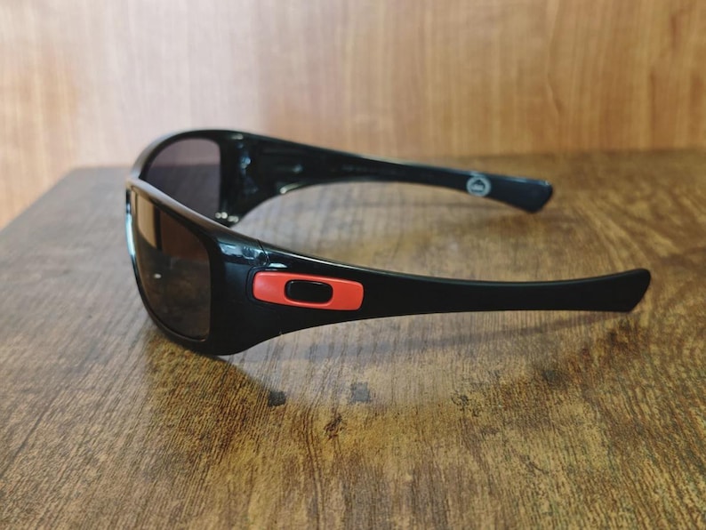 K&ouml;nnte beinhalten: Schwarze Sonnenbrille mit gl&auml;nzendem Finish und dunklen Gl&auml;sern. Die Sonnenbrille hat einen roten Akzent am Rahmen und ist vor einem Holzgrund platziert. Das Design ist sportlich und elegant, geeignet f&uuml;r Outdoor-Aktivit&auml;ten.