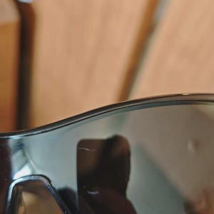 May include: Close-up of black sunglasses with dark tinted lenses. The sunglasses have a sleek, modern design with a subtle curve at the top. The lenses reflect the surrounding environment.