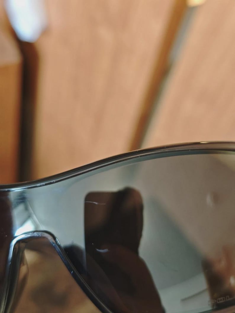 May include: Close-up of black sunglasses with dark tinted lenses. The sunglasses have a sleek, modern design with a subtle curve at the top. The lenses reflect the surrounding environment.
