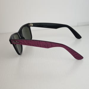 Puede incluir: Gafas de sol con lentes de color verde oscuro y montura negra. Las patillas exteriores presentan un acabado texturizado de color morado intenso con un patr&oacute;n sutil. El logotipo de Ray-Ban es visible en letras plateadas cerca de la bisagra.
