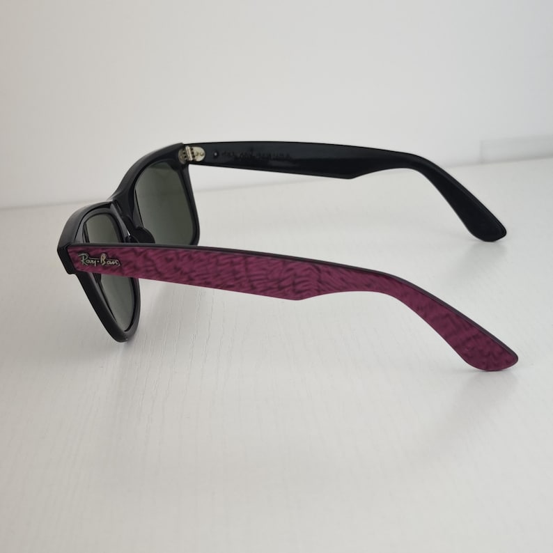 Puede incluir: Gafas de sol con lentes de color verde oscuro y montura negra. Las patillas exteriores presentan un acabado texturizado de color morado intenso con un patr&oacute;n sutil. El logotipo de Ray-Ban es visible en letras plateadas cerca de la bisagra.