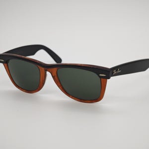 May include: Sunglasses with a classic design. The frames are a combination of black and brown, with dark green lenses. The Ray-Ban logo is visible on the temple.