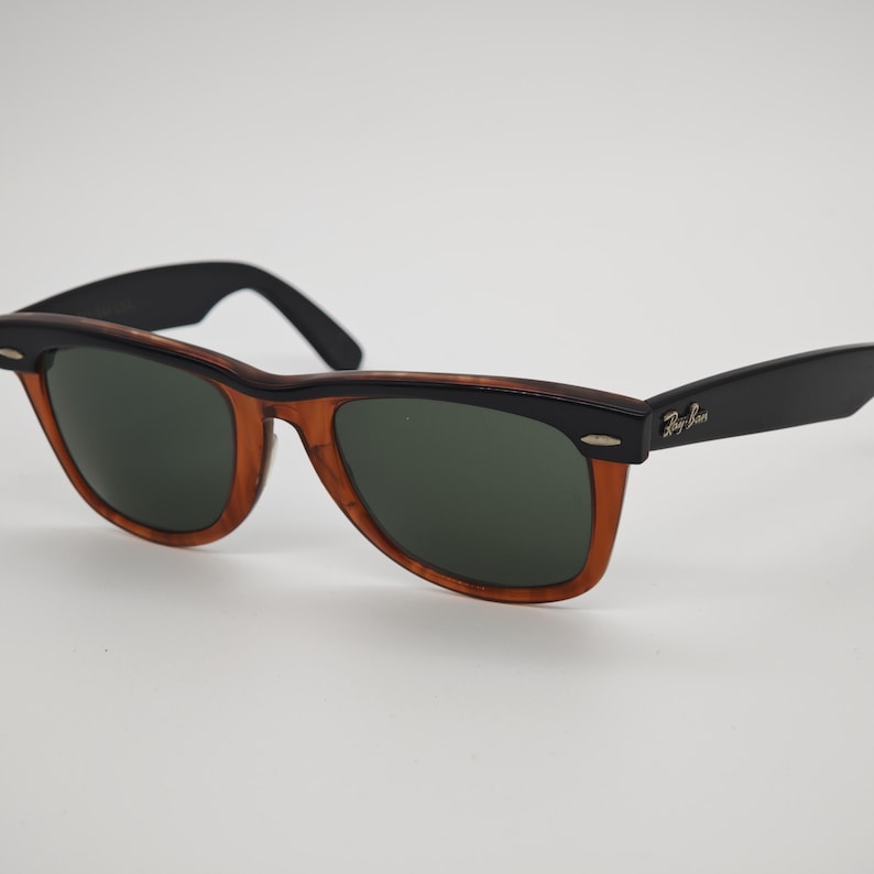 May include: Sunglasses with a classic design. The frames are a combination of black and brown, with dark green lenses. The Ray-Ban logo is visible on the temple.