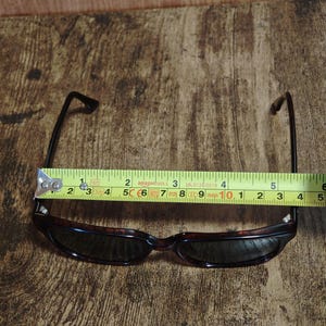 May include: Sunglasses with a tortoiseshell frame and dark lenses. The glasses are measured with a tape measure, showing a width of approximately 5 inches. The arms are black and extend from the frame.