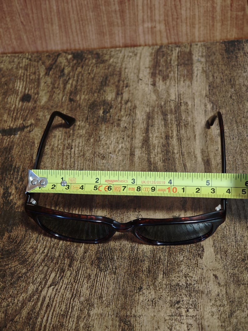 May include: Sunglasses with a tortoiseshell frame and dark lenses. The glasses are measured with a tape measure, showing a width of approximately 5 inches. The arms are black and extend from the frame.